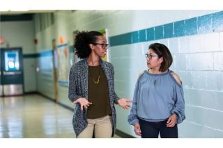 Two multiracial women walking and talking in a school hallway. The woman on the left is mixed race Hispanic and African-American, in her 30s. The person on the right is a Filipino woman in her 40s.